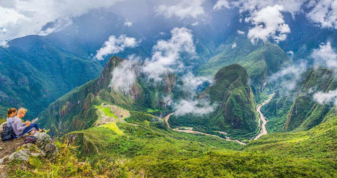 Hiking Macchu Picchu A couple sits dangling their feet over a cliff as they gaze over lush green mountains enclosed in clouds