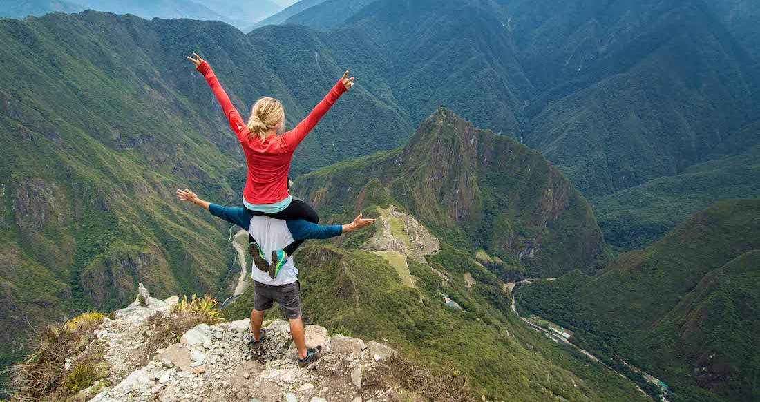Macchu Picchu Peru A women giving the peace sign sits on the shoulders of a man as they overlook a lush mountain range