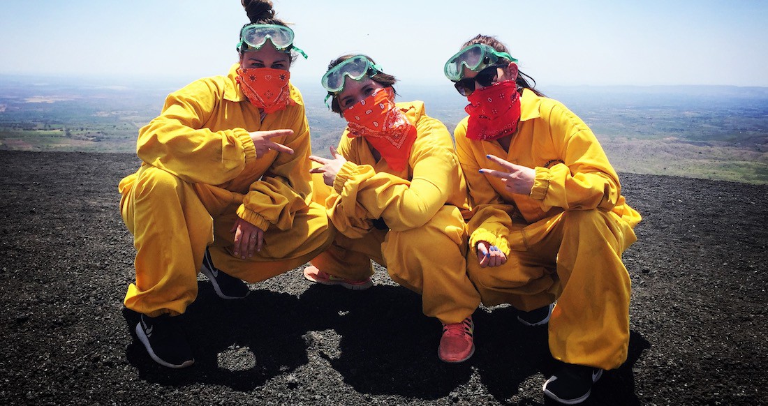 Volcano Boarding Three girls in yellow jumpsuits, green goggles, and orange handkerchiefs kneel on black volcanic sand on top of a steep hill