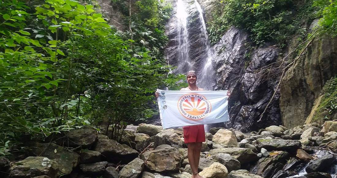 Waterfall Colombia A man stands in front of a waterfall holding a blue flag that says Free and Easy Traveler on it.